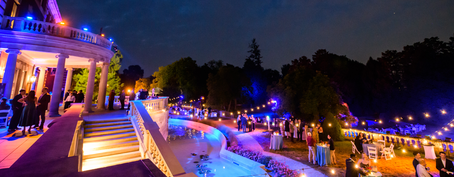 FDU Forward outdoor evening event at a large building with columns and a terrace, where groups of people socialize around tables near a lit pool and garden, with string lights and colorful lighting illuminating the scene.