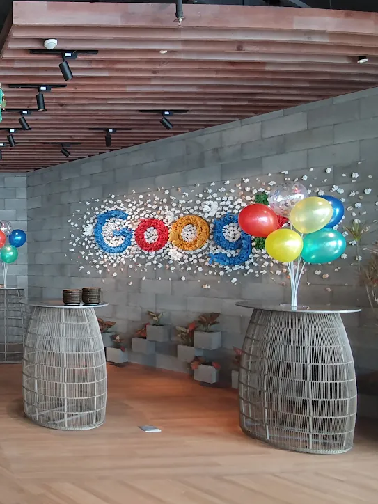 Um escritório do Google decorado para uma comemoração. Há arranjos de balões coloridos nas mesas de trabalho e grandes guarda-chuvas de seda estão pendurados no teto.