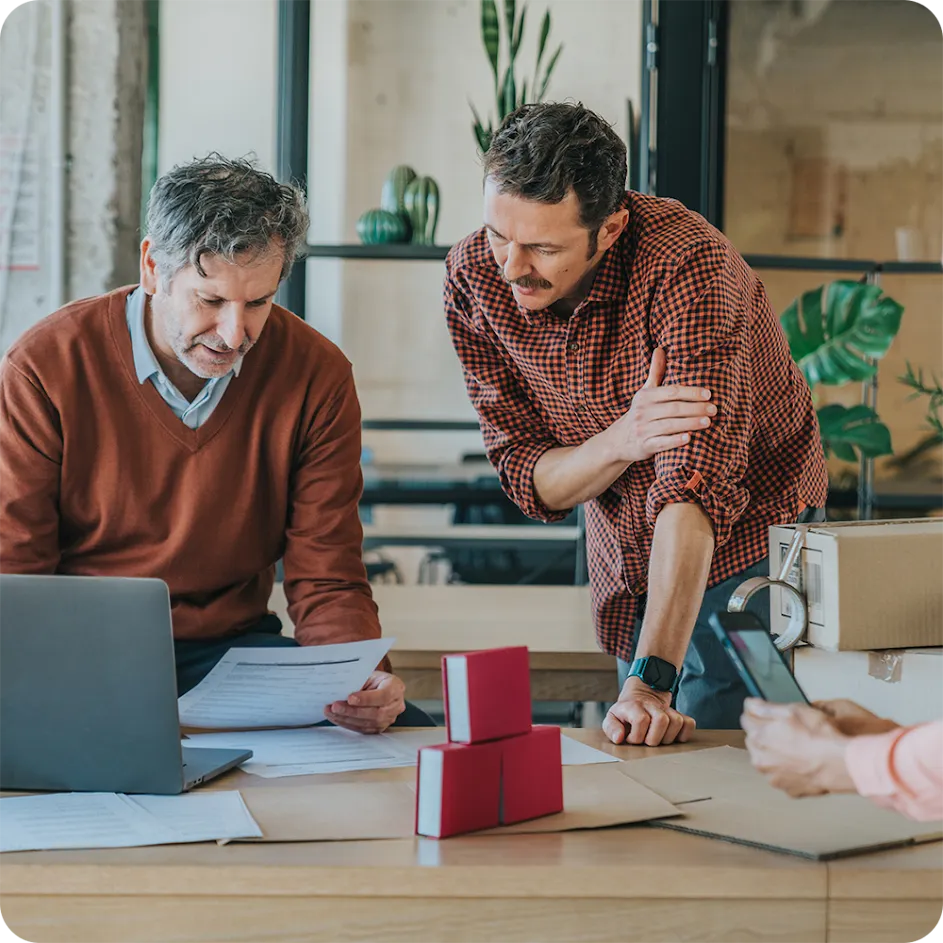 Photograph of two men in an office setting full of plants and natural light, as they review paper documents on a shared desk space where we also see an open laptop, some small red boxes, and another coworker’s phone. 